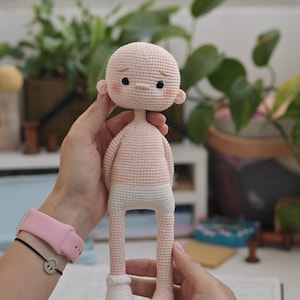 May include: A handmade crochet doll with a pale peach-colored head and torso, featuring small black eyes, embroidered eyebrows, and pink blush on the cheeks. The doll has white crocheted underwear and one white sock on its right foot, with a slender build.