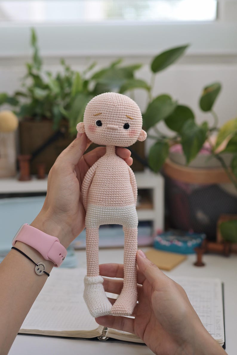 May include: A handmade crochet doll with a pale peach-colored head and torso, featuring small black eyes, embroidered eyebrows, and pink blush on the cheeks. The doll has white crocheted underwear and one white sock on its right foot, with a slender build.
