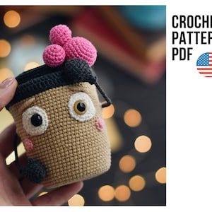Puede incluir: Patrón de crochet PDF para una taza de crochet marrón con una tapa de crochet negra y pompones rosas. La taza tiene una cara con ojos blancos, pupilas negras y mejillas rosas.