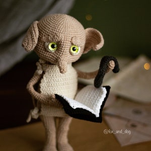 Puede incluir: Un mu&ntilde;eco Dobby de crochet de la serie Harry Potter, sosteniendo un libro y una varita m&aacute;gica. El mu&ntilde;eco lleva un vestido blanco y tiene grandes ojos verdes.