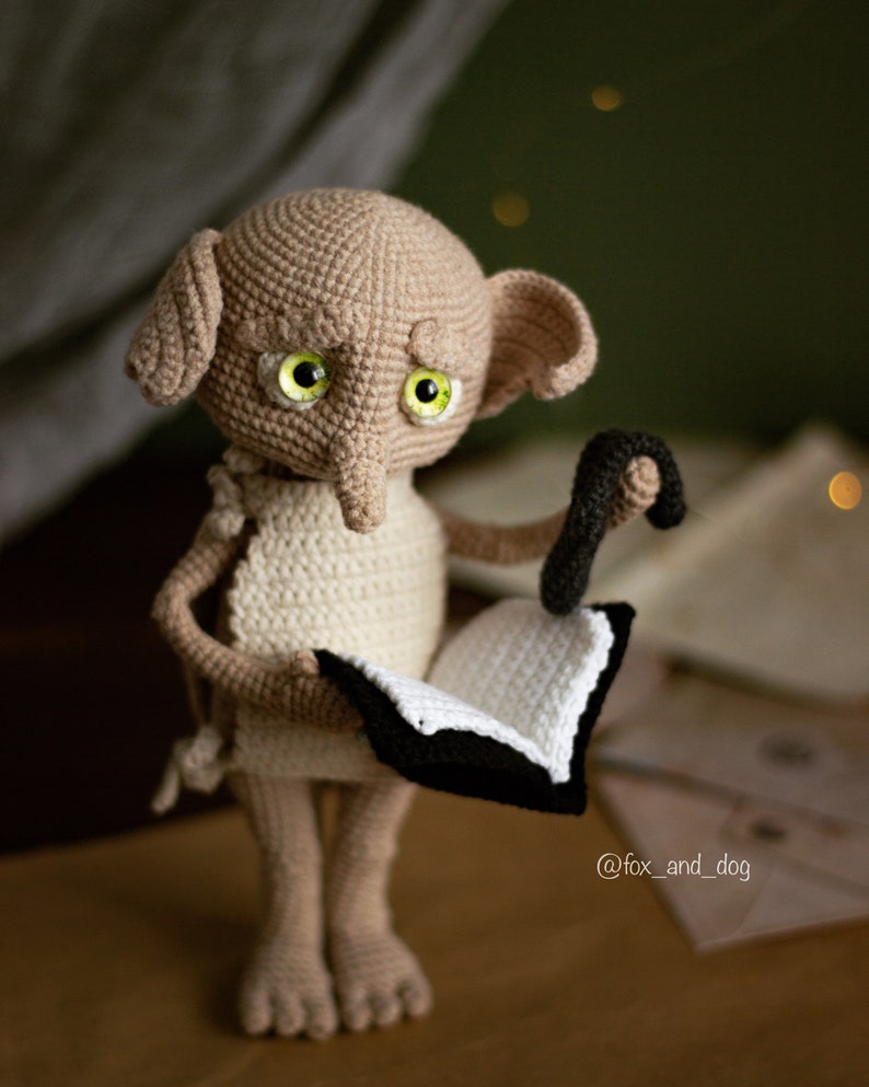 Puede incluir: Un mu&ntilde;eco Dobby de crochet de la serie Harry Potter, sosteniendo un libro y una varita m&aacute;gica. El mu&ntilde;eco lleva un vestido blanco y tiene grandes ojos verdes.