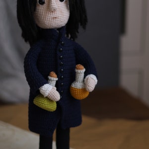 May include: Crocheted doll of a fictional character with long black hair, wearing a dark blue coat and holding three small bottles. The doll is standing on a brown surface.