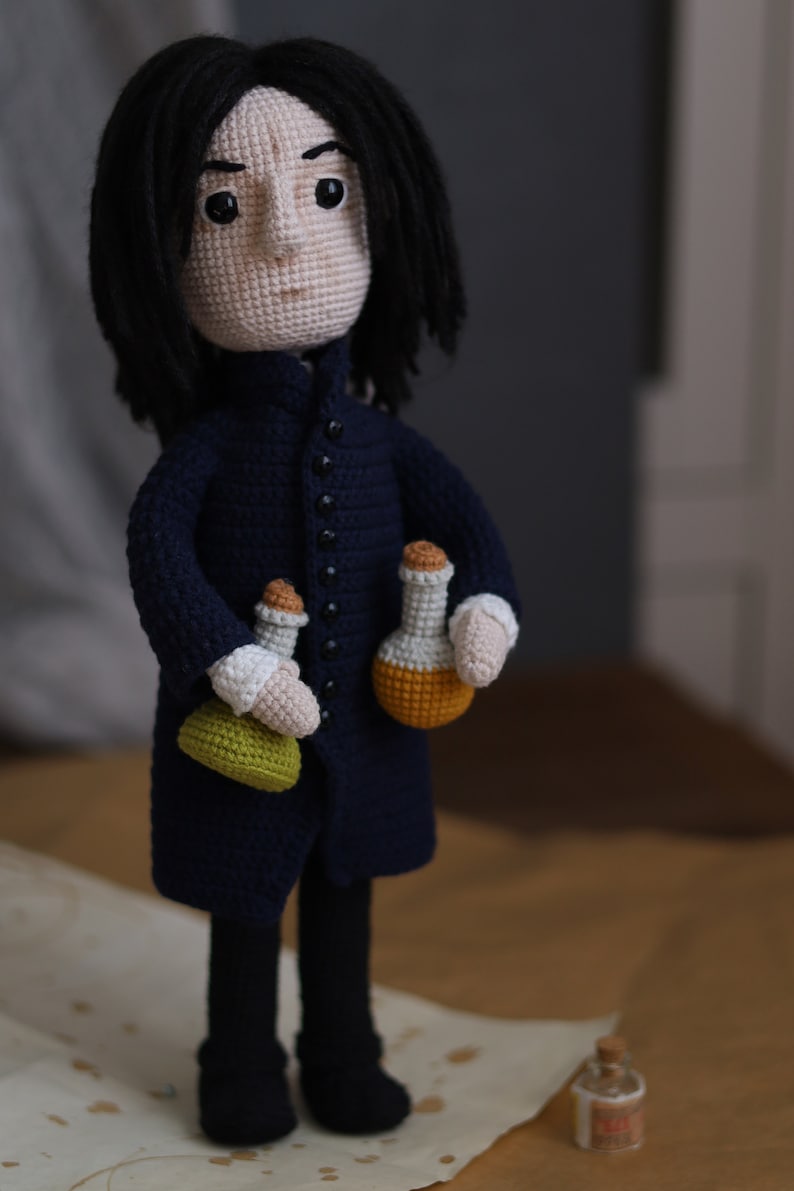 May include: Crocheted doll of a fictional character with long black hair, wearing a dark blue coat and holding three small bottles. The doll is standing on a brown surface.