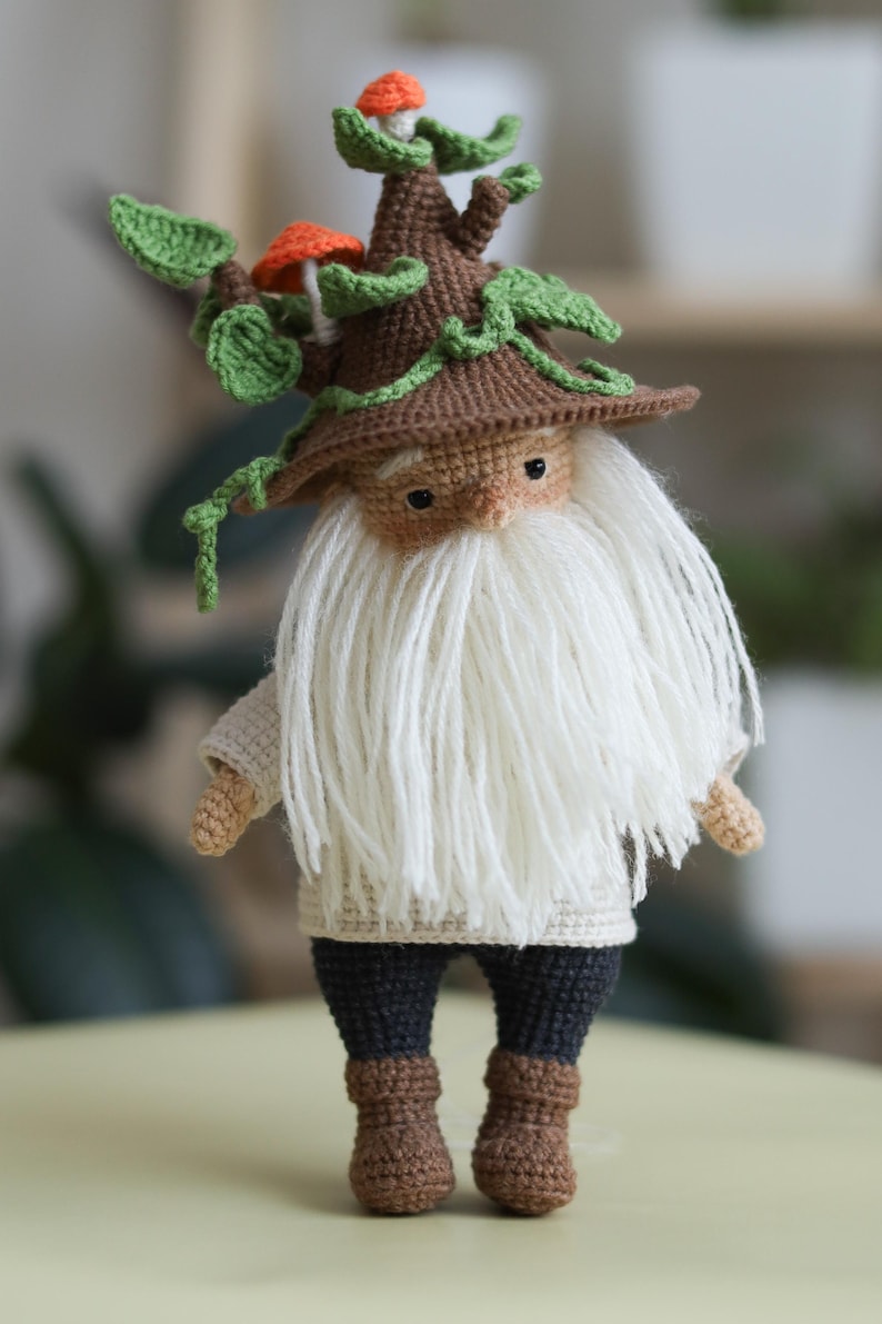 May include: A crocheted gnome doll wearing a brown hat with green leaves and orange mushrooms. The gnome has a white beard and is wearing a beige sweater, black pants, and brown boots.