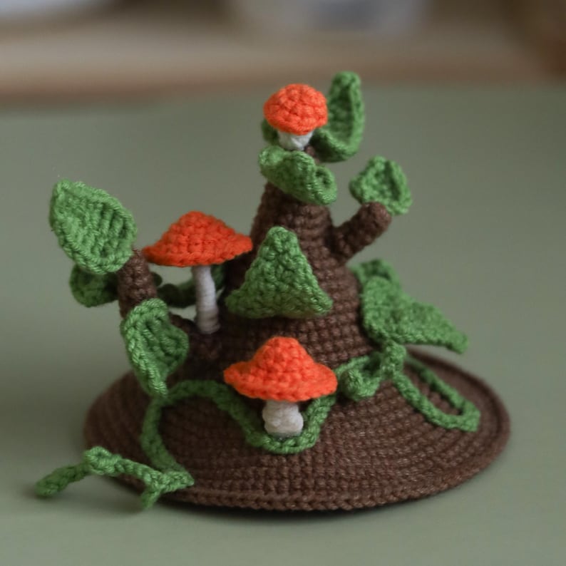 May include: Crocheted mushroom tree with orange and white mushrooms and green leaves. The tree is brown and has a base that looks like a tree stump.