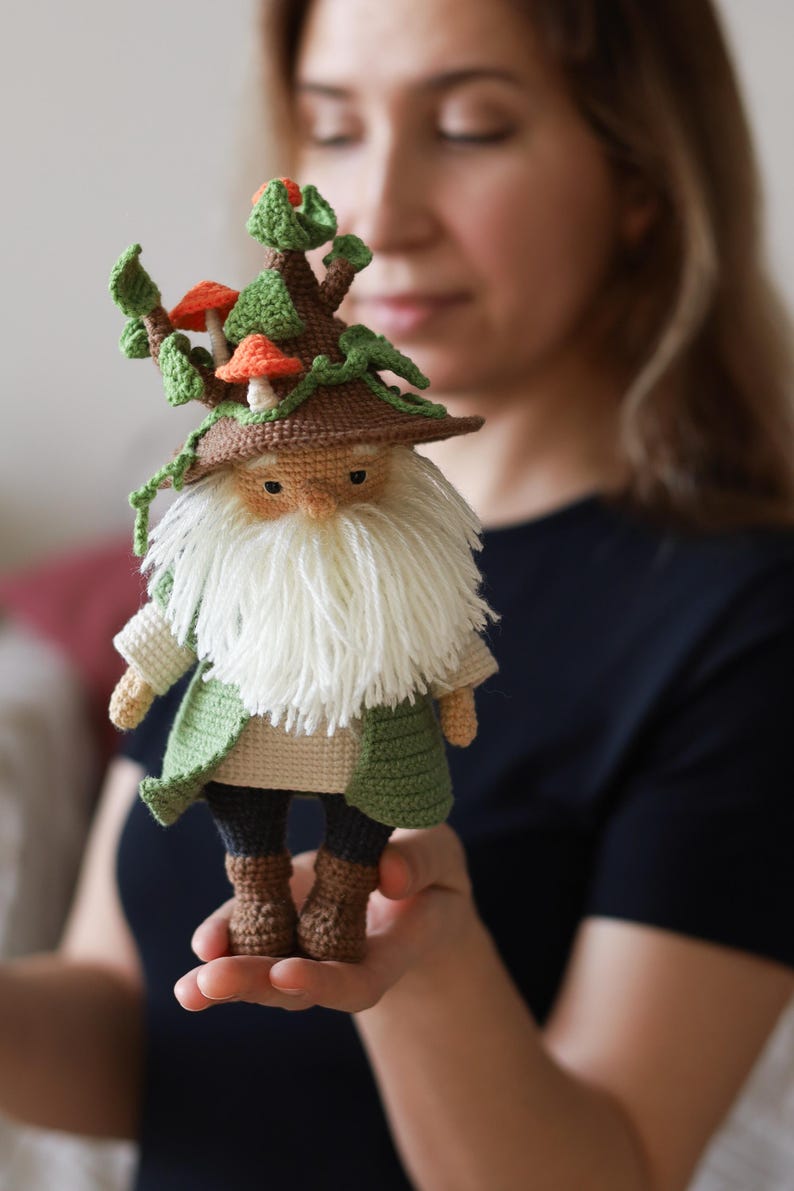 May include: A crocheted gnome doll wearing a brown hat with green leaves and orange mushrooms. The gnome has a white beard and is wearing a green and brown outfit.