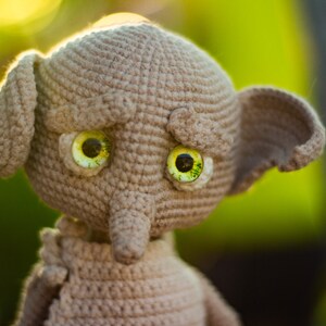 Puede incluir: Un juguete de peluche tejido a crochet con un cuerpo marr&oacute;n y grandes ojos verdes. El juguete tiene una nariz larga y puntiaguda y orejas grandes.