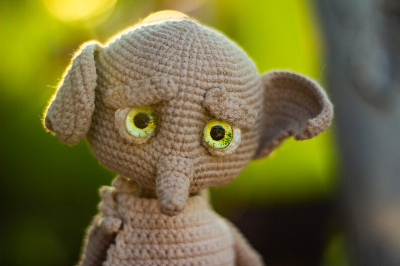 Puede incluir: Un juguete de peluche tejido a crochet con un cuerpo marr&oacute;n y grandes ojos verdes. El juguete tiene una nariz larga y puntiaguda y orejas grandes.