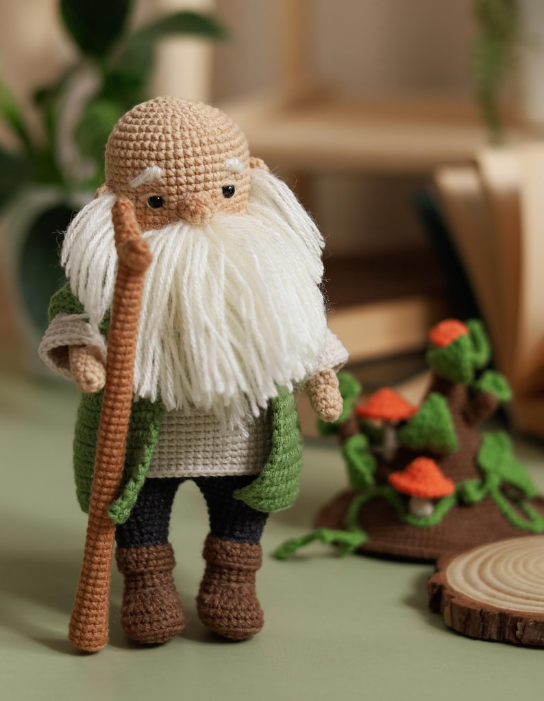 May include: A crocheted gnome doll with a long white beard, wearing a green jacket and brown pants, holding a wooden stick. The gnome is standing on a green surface with a crocheted mushroom and tree stump in the background.