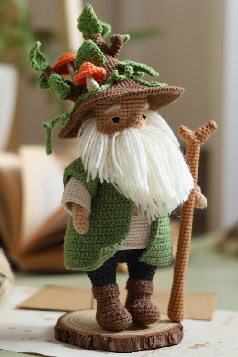 May include: A crocheted gnome doll with a brown hat topped with green leaves and orange mushrooms. The gnome is wearing a green jacket, brown pants, and brown boots. The gnome is holding a brown stick.