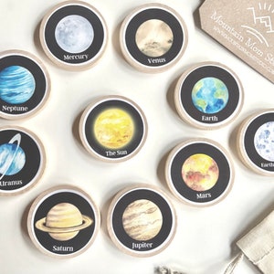 May include: A set of ten round wooden discs with black backgrounds and watercolour illustrations of the planets in our solar system, including the sun, Earth's moon, and the names of each celestial body.