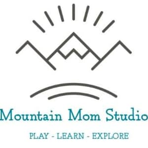 以下が含まれることがあります： 遊び、学び、探検の機会を提供するMountain Mom Studioのロゴ。ロゴは、太陽が輝くスタイリッシュな山脈を特徴としています。「Mountain Mom Studio」のテキストは青色のフォントで、「PLAY - LEARN - EXPLORE」のテキストはより小さく、灰色のフォントです。