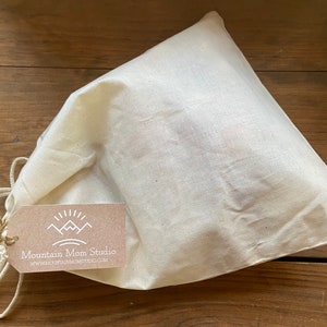 以下が含まれることがあります： 白い綿の巾着袋で、茶色のタグには「Mountain Mom Studio」と「www.mountainmomstudio.com」と書かれています。