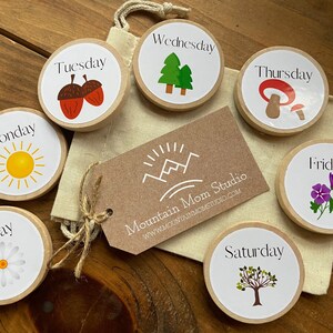May include: A set of seven wooden day of the week circles with illustrations and text. The circles are in a bag with a brown tag that says "Mountain Mom Studio" and a website address. The illustrations include a sun, acorns, trees, a mushroom, a flower, and a tree.