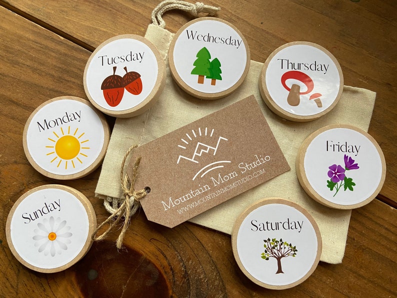 May include: A set of seven wooden day of the week circles with illustrations and text. The circles are in a bag with a brown tag that says "Mountain Mom Studio" and a website address. The illustrations include a sun, acorns, trees, a mushroom, a flower, and a tree.