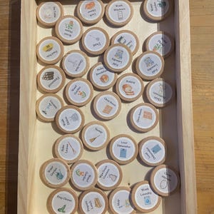 May include: A set of wooden tokens with illustrations and text representing household chores. The tokens are arranged in a wooden tray. Some of the chores include: Cook Dinner, Set Table, Dust, Water Plants, Fold Laundry, Baking, Clean Bathroom, Clean Room, Weed Garden, Load Dishwasher, Clean Mirror, Wash Laundry, Wipe Down Counters, and Prep Dinner.