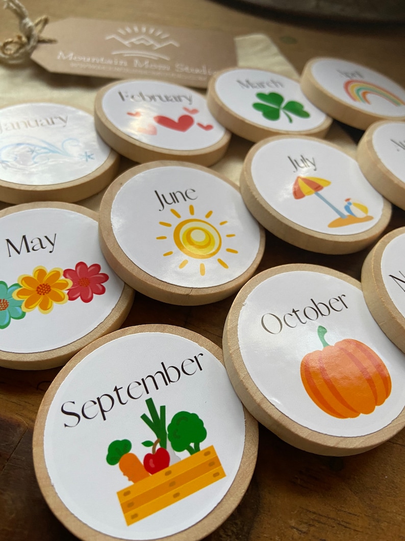 May include: Twelve wooden circles with a different month of the year and a corresponding image on each. The months are January, February, March, April, May, June, July, August, September, October, November, and December. The images include a snowflake, a heart, a shamrock, a rainbow, a flower, a sun, an umbrella, a star, a vegetable garden, a pumpkin, a turkey, and a snowman.