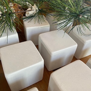 Pode incluir: Uma coleção de blocos quadrados brancos, possivelmente para fins decorativos, dispostos sobre uma superfície de madeira. Alguns blocos são adornados com raminhos de agulhas de pinheiro verdes e pequenas pinhas, sugerindo um tema festivo.