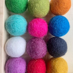 Puede incluir: Doce bolas de fieltro de colores en varios tonos de verde, azul, rosa, morado, rojo, naranja, amarillo, blanco y negro. Las bolas están dispuestas en tres filas de cuatro, con cada fila presentando una combinación de colores diferente.
