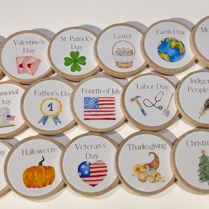 May include: A set of 16 round wooden tokens with illustrations and text representing different holidays throughout the year.  The tokens feature images such as fireworks, hearts, shamrocks, Easter eggs, a globe, a flower, a tombstone, a number one, an American flag, a hammer, a stethoscope, a feather, a pumpkin, a heart with stars, a cornucopia, and a Christmas tree.