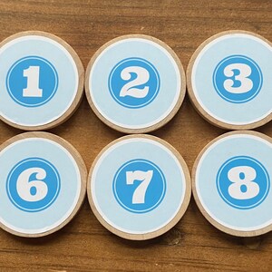 Pode incluir: Dez peças redondas de madeira com centros azuis e números brancos de 0 a 9.