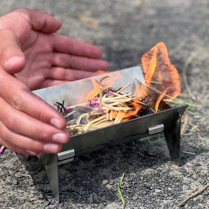 Peut inclure: Un petit brasero portable en métal argenté à quatre pieds, contenant un tas d'allumettes en bois à bouts roses en train de brûler. Les flammes s'élèvent du centre de la structure métallique, qui est posée sur une surface de gravier.