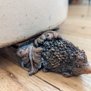 Peut inclure: Gros plan d'un pot en céramique décoratif avec un pied en forme de hérisson. Le pied est en métal brun avec une surface texturée. Le pot est blanc avec un bord brun.