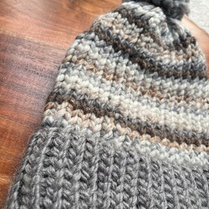 Pu&ograve; includere: Un berretto di lana a maglia a righe grigie, marroni e bianche con un pompon grigio soffice in cima.