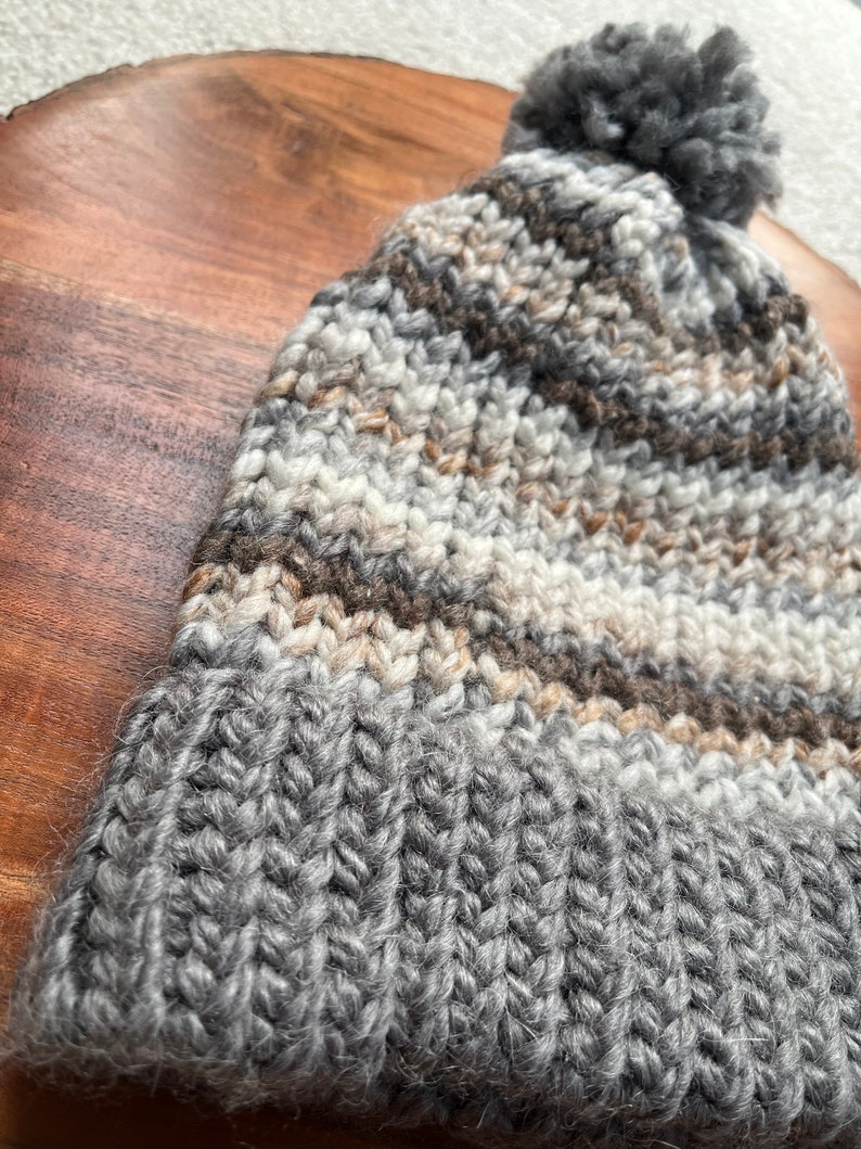 Pu&ograve; includere: Un berretto di lana a maglia a righe grigie, marroni e bianche con un pompon grigio soffice in cima.