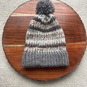 Pu&ograve; includere: Un berretto di lana a righe grigie e marroni con un pompon grigio in cima.
