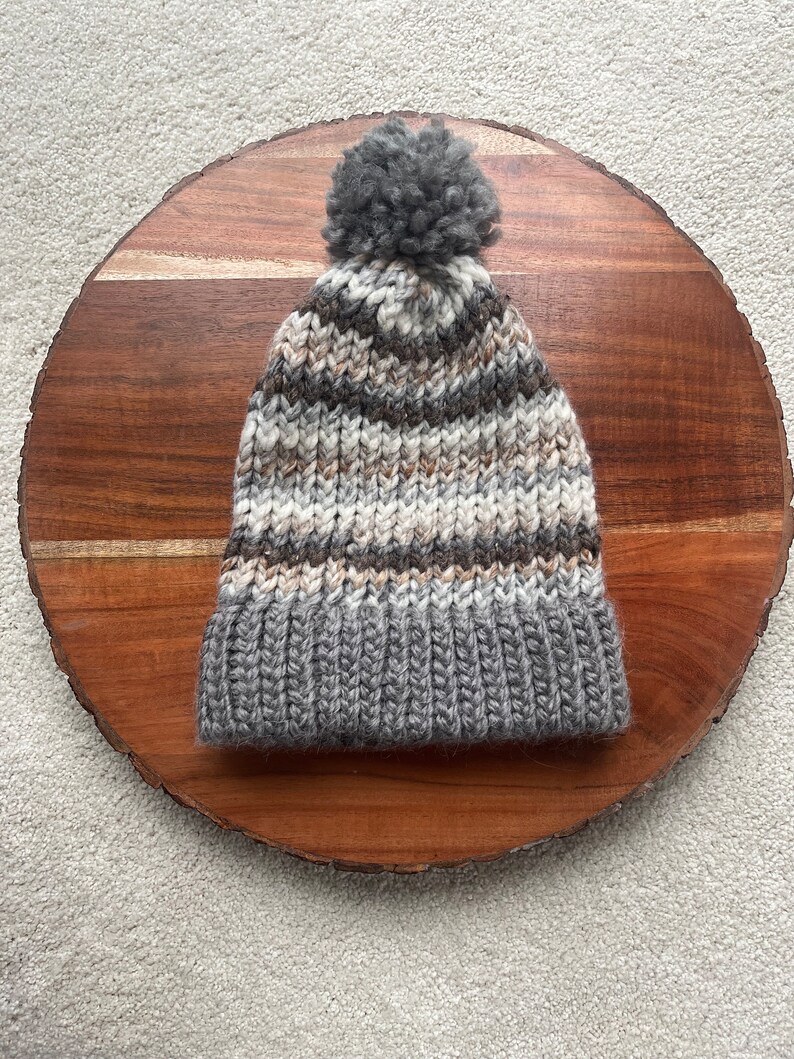 Pu&ograve; includere: Un berretto di lana a righe grigie e marroni con un pompon grigio in cima.