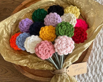 Può includere: Un bouquet di fiori all'uncinetto in vari colori, tra cui rosso, arancione, giallo, verde, blu, viola e rosa. I fiori sono disposti in una ciotola di legno e avvolti in carta velina dorata. Un piccolo cartellino con il testo "hello gorgeous" è attaccato al bouquet.