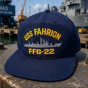 May include: A navy blue baseball cap featuring an embroidered patch on the front. The patch displays a grey ship silhouette with the yellow text USS FAHRION above and FFG-22 below. The cap has a structured crown and a flat brim.