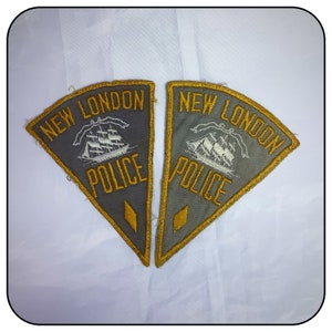 May include: Two gray triangular patches with gold trim. Each patch has the text "NEW LONDON POLICE" in gold lettering and a white embroidered image of a ship.