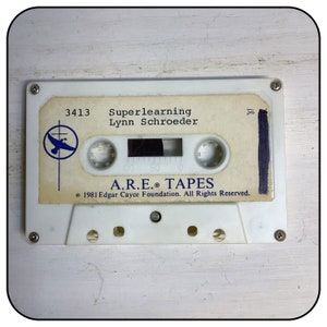 May include: A white cassette tape featuring a label with the text 3413 Superlearning Lynn Schroeder, a blue bird logo, and A.R.E. TAPES 1981 Edgar Cayce Foundation. The cassette has four visible screws and a central window showing dark magnetic tape.