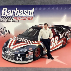 May include: A red, white, and blue race car with the number 38 and the words "Barbasol" on the side. A man in a white shirt and black pants stands next to the car. The car is on a red and white background with stars.