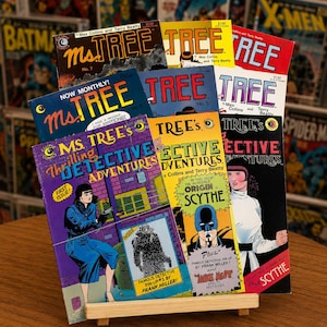 May include: A collection of Ms. Tree comic books displayed on a wooden stand. The covers feature colorful illustrations, bold typography, and titles such as Ms. Tree, Thrilling Detective Adventures, and The Origin of Scythe, with various issue numbers.