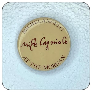 May include: A beige button with the text "Michelangelo" and "Mi & Cagnio Co. At The Morgan" in red lettering.