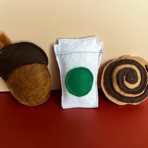 May include: Three felt toys consisting of a brown acorn with a textured cap, a white cup with a green circular emblem, and a tan circular pastry with a dark brown spiral design. Each item is crafted from soft fabric with visible stitching.