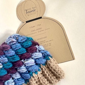 May include: A colourful crocheted beanie hat with a brown tag that says "Messy Bun Beanie". The tag also includes care instructions for the hat.