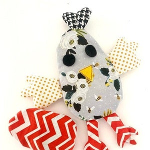 May include: A handmade plush toy shaped like a bird with a gray body, white flowers, and yellow bees. The bird has red and white striped legs and wings made of yellow polka dot fabric and red and white chevron fabric.