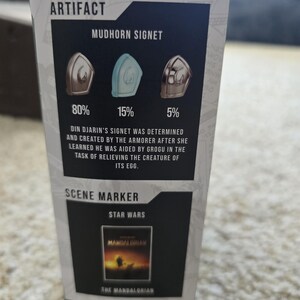 Puede incluir: Una secci&oacute;n de una caja de producto de Star Wars con el texto "ARTIFACT" y "MUDHORN SIGNET". Muestra tres dise&ntilde;os de signet con porcentajes. El texto debajo describe el origen del signet. Una secci&oacute;n "SCENE MARKER" muestra una imagen de "The Mandalorian".