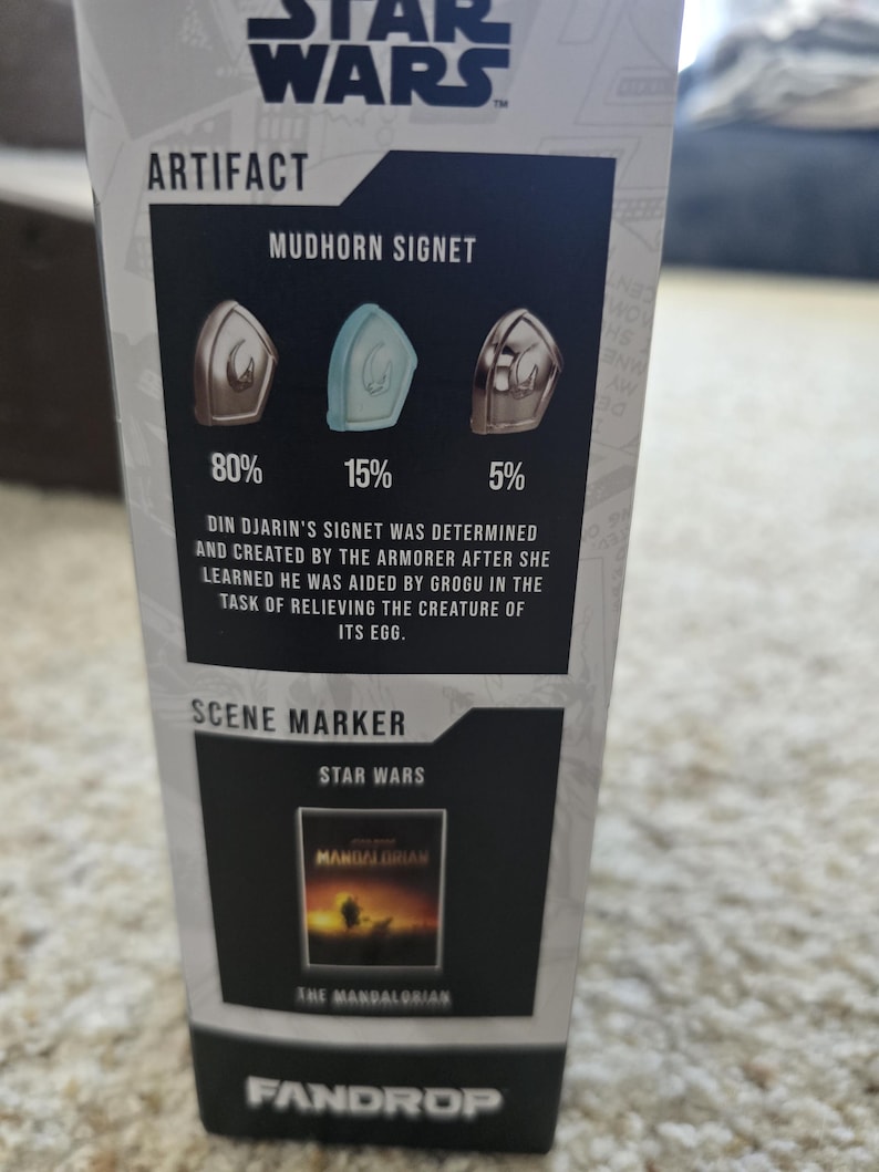 Puede incluir: Una secci&oacute;n de una caja de producto de Star Wars con el texto "ARTIFACT" y "MUDHORN SIGNET". Muestra tres dise&ntilde;os de signet con porcentajes. El texto debajo describe el origen del signet. Una secci&oacute;n "SCENE MARKER" muestra una imagen de "The Mandalorian".