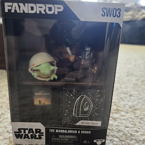 Puede incluir: Un set de caja Fandrop de Star Wars con figuras de The Mandalorian y Grogu. La caja incluye una peque&ntilde;a figura de Grogu con casco, una figura de Mandaloriano y un peque&ntilde;o libro. La caja tambi&eacute;n tiene el logo de Star Wars y el texto "Artifact Inside".