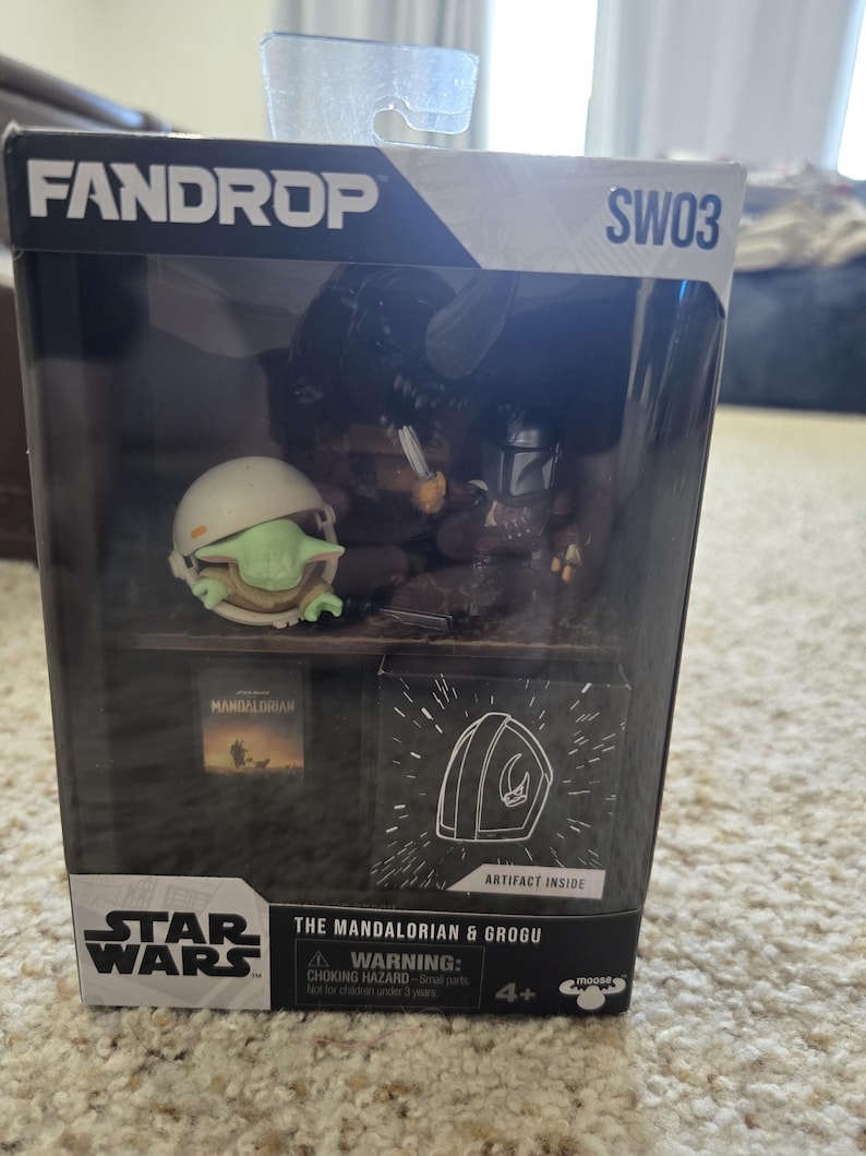 Puede incluir: Un set de caja Fandrop de Star Wars con figuras de The Mandalorian y Grogu. La caja incluye una peque&ntilde;a figura de Grogu con casco, una figura de Mandaloriano y un peque&ntilde;o libro. La caja tambi&eacute;n tiene el logo de Star Wars y el texto "Artifact Inside".