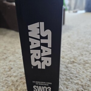 Puede incluir: Una caja negra con la palabra "FANDROP" en la parte superior. Las palabras "STAR WARS" est&aacute;n impresas en letras blancas grandes. Debajo, el texto dice "THE MANDALORIAN & GROGU THE MANDALORIAN" y "SW03".