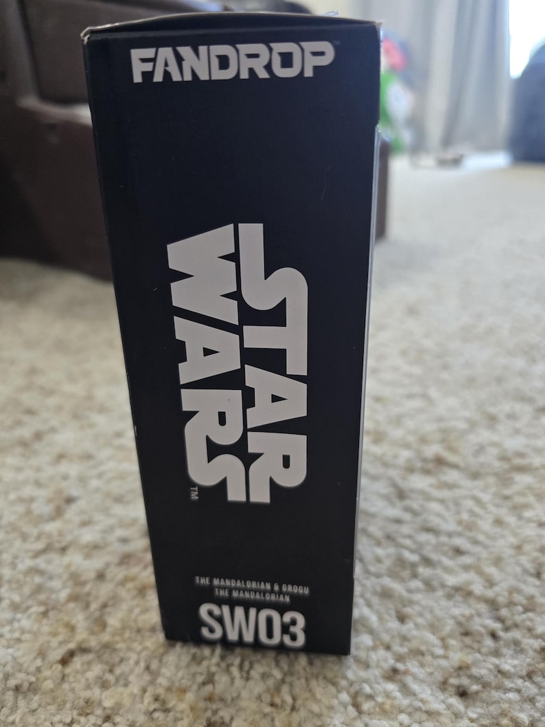 Puede incluir: Una caja negra con la palabra "FANDROP" en la parte superior. Las palabras "STAR WARS" est&aacute;n impresas en letras blancas grandes. Debajo, el texto dice "THE MANDALORIAN & GROGU THE MANDALORIAN" y "SW03".