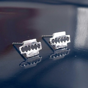 May include: A pair of silver-colored stud earrings. Each earring features a stylized, symmetrical design with a rectangular shape and a series of curved cutouts along the bottom edge. The earrings are set against a dark blue, reflective surface.