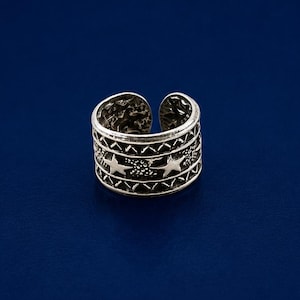 May include: A silver-colored adjustable ring with a detailed design. The ring features a band with star and geometric patterns. The ring is set against a dark blue background.