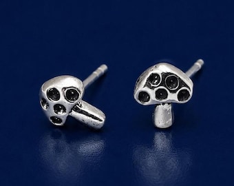 May include: A pair of silver-toned stud earrings shaped like mushrooms. Each earring features a rounded cap with recessed circular details and a short, smooth stem. The metal has a polished finish with darkened accents in the crevices.